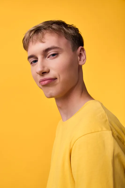 Young man wearing a yellow shirt stands against a bright yellow backdrop, presenting casual fashion with a confident expression in a clean studio setting, close up head and shoulders shot