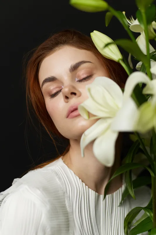 A serene model with fair skin and reddish brown hair poses gracefully with white lilies, dressed in a white blouse, conveying calmness and beauty