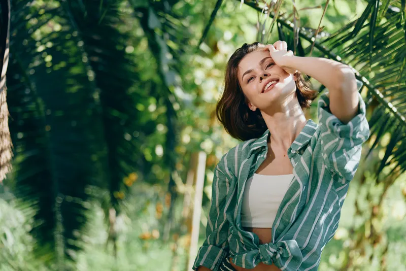 Bright outdoor portrait of a smiling woman in a green striped shirt, standing in a sunlit garden.