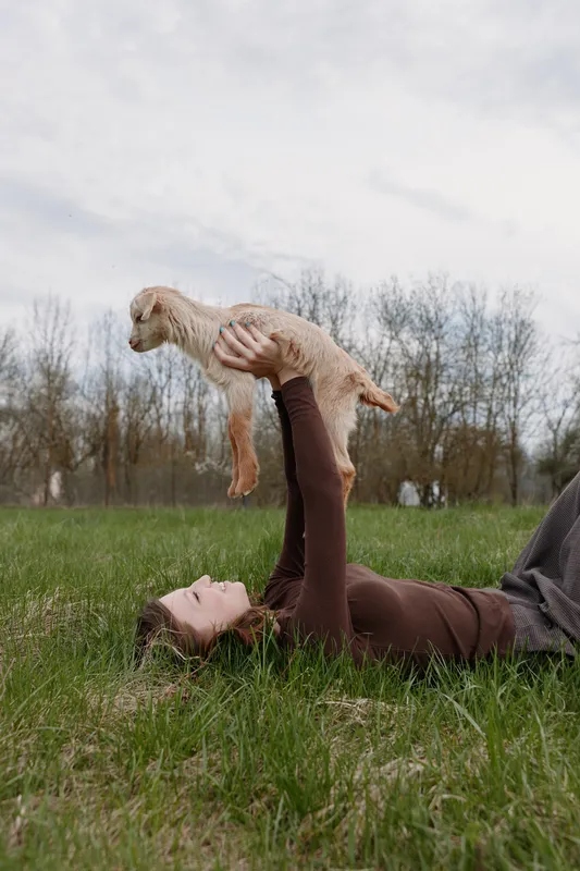 Joyful outdoor moment with a woman lying on grass, lifting a curious goat kid above her, sunlit scene, relaxed posture, candid lifestyle amid natural greens
