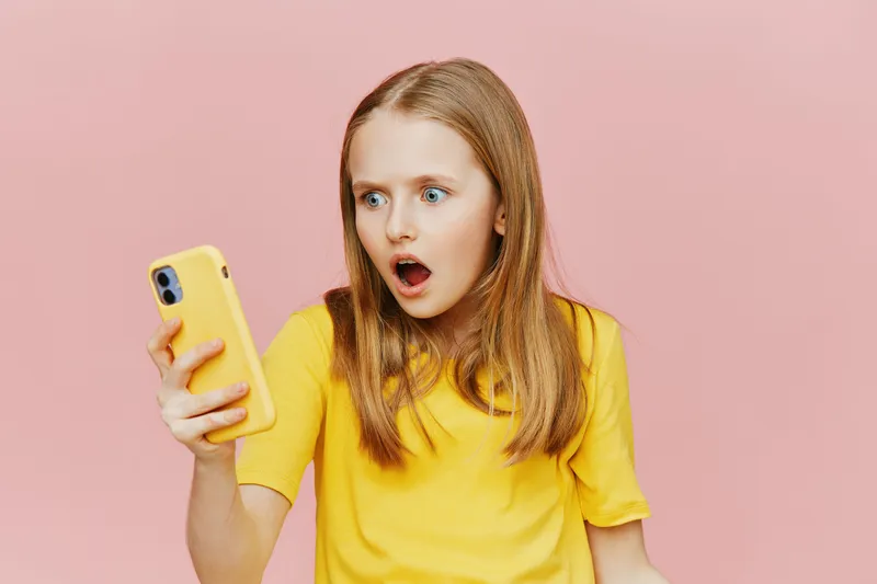 Surprised girl in yellow t shirt looking at smartphone against pink background, expressing shock and excitement