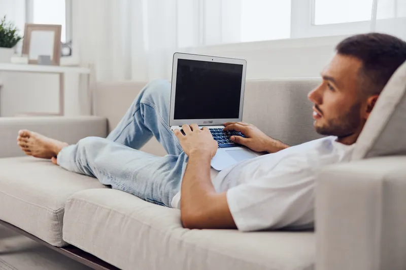 Young man relaxing at home on a couch, working with a laptop, displaying a casual and modern lifestyle, perfect for home office themes in 2025 and 2026.