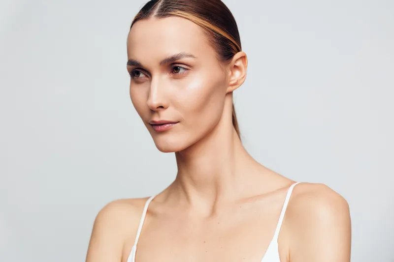 A young woman with smooth skin and slicked-back hair looks to the side against a plain light background. The image highlights natural beauty and skincare concepts.