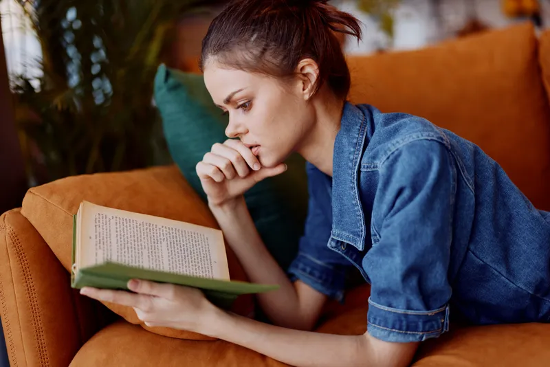 Женщина отдыхает и читает книгу, устроившись на удобном диване в уютной гостиной; на заднем плане — зелёное растение