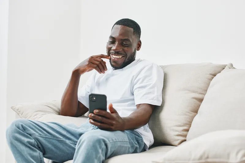 Happy young Black man sitting on a couch and using a smartphone, wearing casual white t shirt and jeans, indoors in a bright modern room