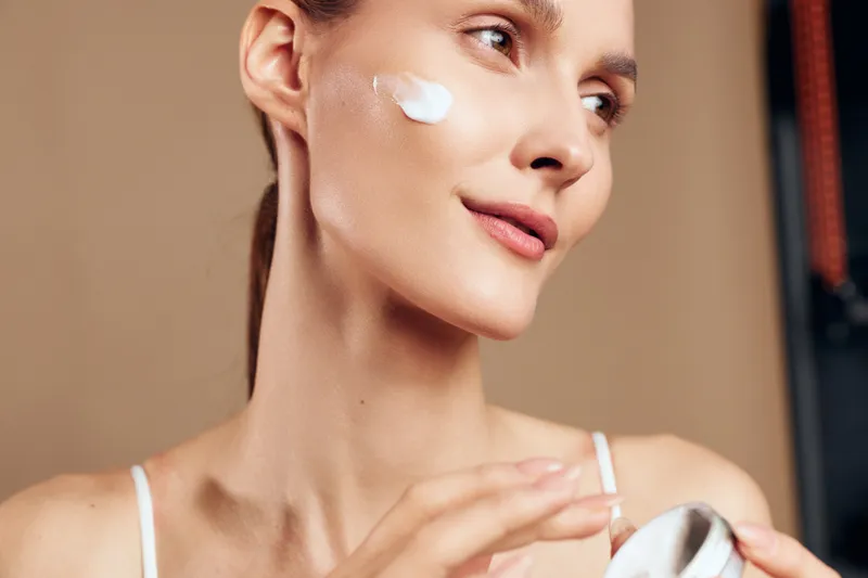 Young woman applies moisturizing cream on her cheek with gentle smile close up natural skincare routine healthy glowing skin beauty concept indoors beige background.