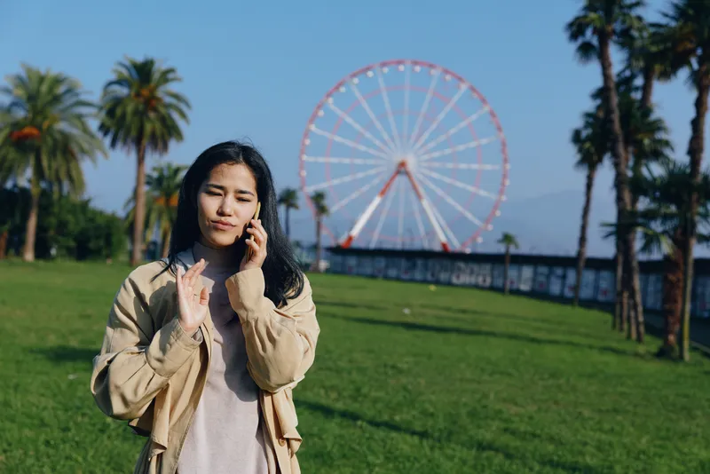 Park scene with a woman on a phone, outdoor setting, greenery, casual jacket, sunny weather, a distant ferris wheel in view, capturing a candid urban leisure moment