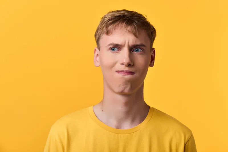 Young man in a yellow shirt against a yellow backdrop shows confusion and curiosity, a facial expression of doubt and hesitation after unexpected news or a tough decision.