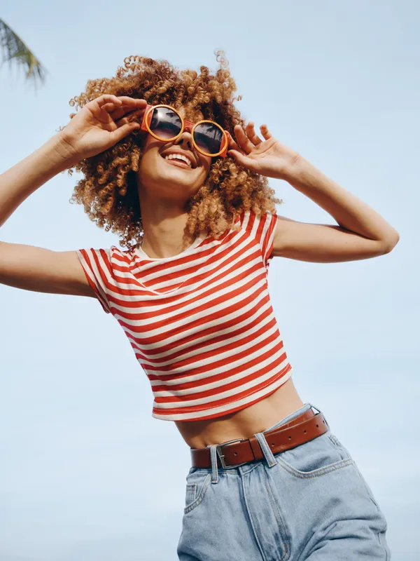 A joyful young woman dances in bright sunlight, wearing a red and white striped crop top, high waisted denim, and round sunglasses, radiating carefree summer energy and fashion flair.