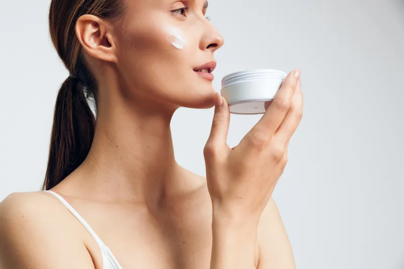 A young woman with clear skin applies white face cream on her cheek holding a jar in hand. The bright background highlights skincare routine and natural beauty concept.