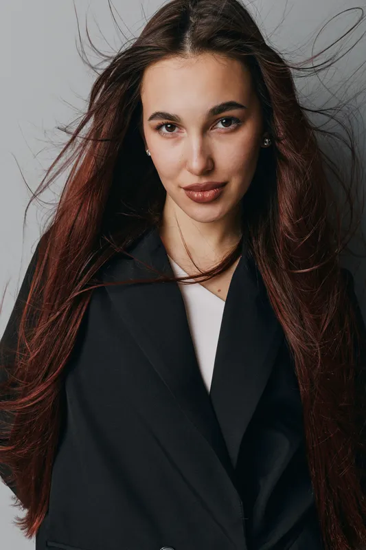 Woman with long hair and confident expression wearing black coat and white top standing against gray background in studio portrait fashion style with natural makeup.