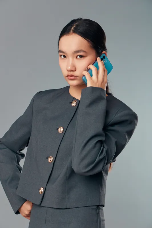 Young woman wearing grey business suit talking on smartphone with serious expression. Portrait of professional female using mobile phone for communication on neutral background.