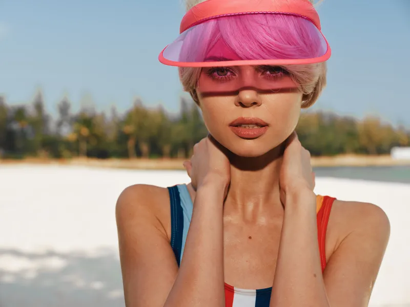 A fashion portrait on a sunny beach shows a woman wearing a pink visor, bold makeup, and a sleeveless swimsuit. Bright light highlights hair and skin for striking contrast.