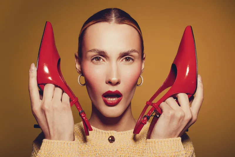 Young woman with slicked hair holds red high heels near face, wearing yellow sweater and hoop earrings. Studio portrait on brown background with fashion and style concept.