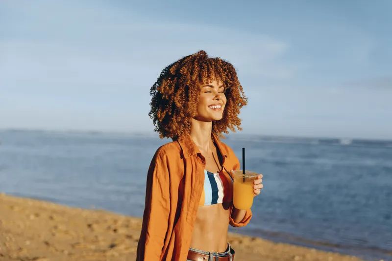 Woman on beach by ocean, curly hair, smiling in warm summer sun, holding a cup, wearing an open jacket and bikini top, enjoying seaside travel lifestyle, outdoors, natural light, relaxed mood