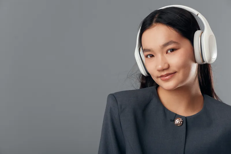 Young woman with long black hair wearing headphones and dark blazer smiling gently on gray background. Portrait, technology, lifestyle and music concept with casual elegance and youth culture.
