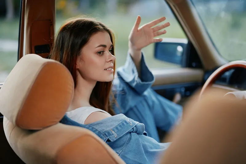 Young woman enjoying a casual moment in a vintage car, dressed in a denim jacket, expressing a relaxed and cheerful emotion against a soft, natural background.