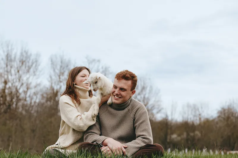 Lifestyle couple outdoors affection with a goat kid in sunlit meadow, relaxed posture, cozy sweaters, natural colors, candid smiles, gentle breeze, peaceful countryside