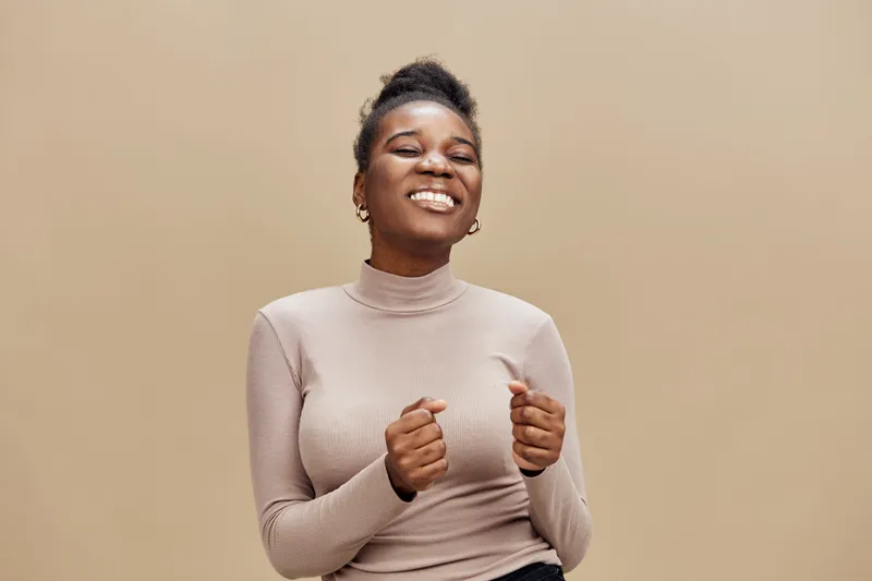 Happy young African woman smiling joyfully in a beige turtleneck on a neutral background, expressing excitement and positivity