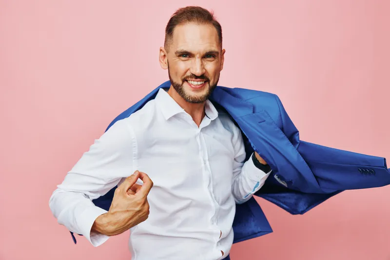 A focused business professional man in a white shirt poses with a blue blazer off his shoulder against an isolated pink background, conveying confidence and determination.