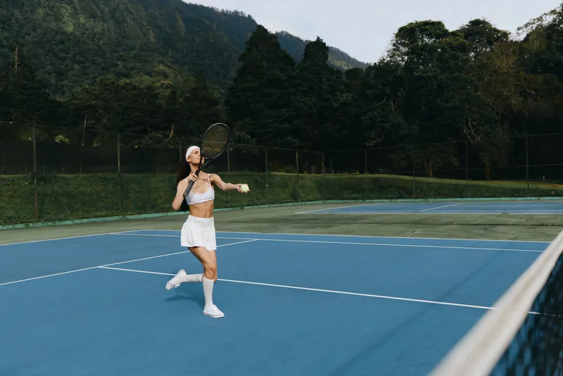 Young athletic woman playing tennis outdoors on a sunny day, wearing a white sports outfit, showcasing excitement and dedication on a vibrant blue court