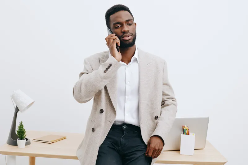 Professional young African American man in business casual attire talking on mobile phone while standing near desk with laptop and office supplies, isolated on plain white background. Office lifestyle