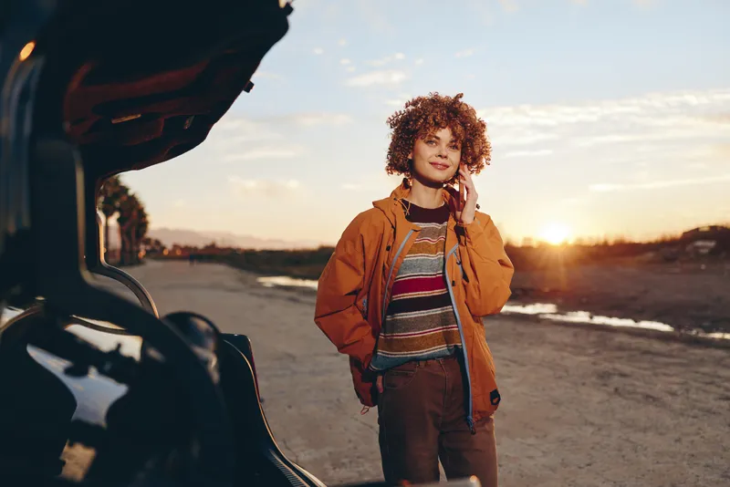 Woman lifestyle at sunset, curly haired woman by a car on a quiet road, warm outdoors travel mood, phone in hand, casual portrait, golden light