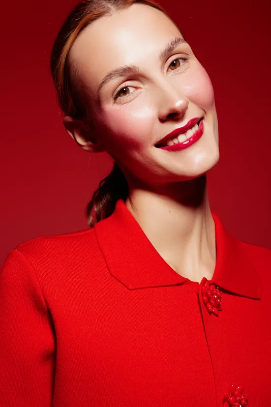 Smiling woman with red hair and bright red lipstick wears red textured jacket against red background. Portrait with soft lighting highlights natural skin and confident expression. Fashion concept.