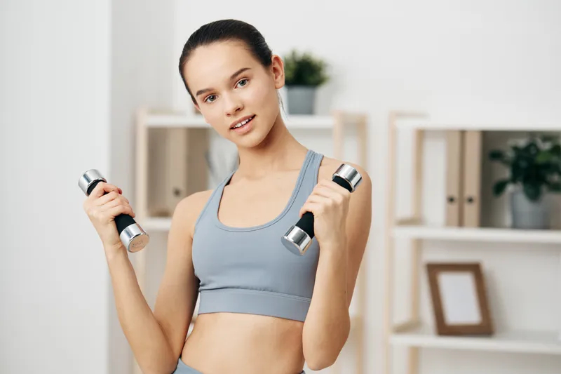 Female fitness enthusiast lifts dumbbells at home in a light, airy room. Athletic wear, focused expression, and a tidy background convey dedication to strength training and a healthy lifestyle.