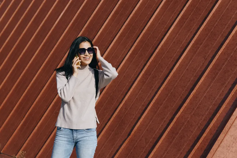 Woman wearing sunglasses and a light sweater stands against a geometric wooden wall, showcasing casual fashion, urban lifestyle, and confident pose for travel, street style, and everyday outdoor