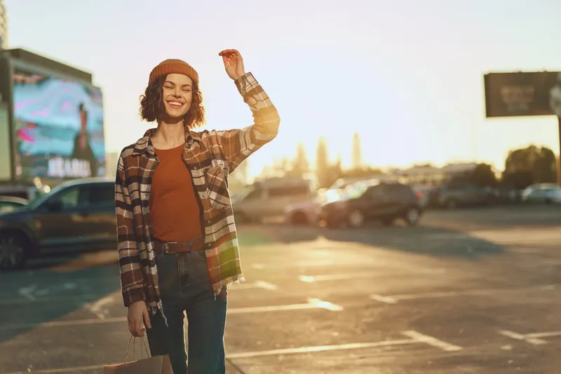 Woman smile shopping in urban sunset parking with bag and casual outfit, candid lifestyle portrait with authenticity and golden hour glow, mindful living and emotional storytelling vibe.