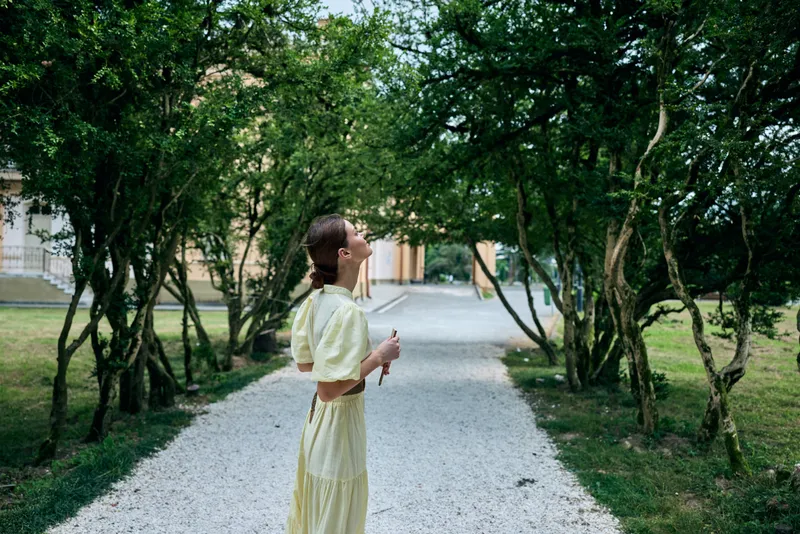 Woman in a light yellow dress stands on a gravel path among lush trees in a sunny park, enjoying a tranquil outdoor moment, breathing fresh air and soaking in nature.