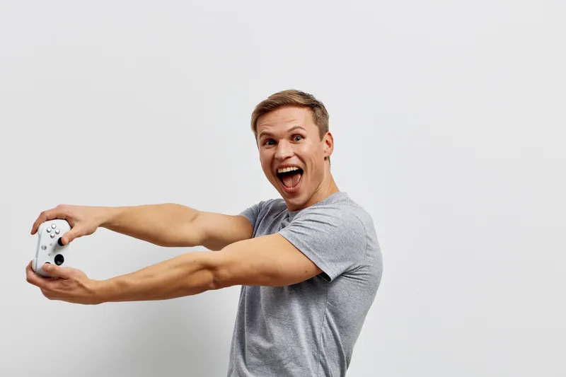 Man taking a selfie with a video game controller, excited and happy expression, casual gray t-shirt, light background, indoor lifestyle shot, entertainment concept.