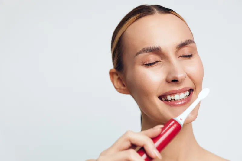 A smiling young woman with closed eyes holds a red electric toothbrush near her mouth in a bright studio. Concept for dental hygiene, oral care, and personal routine.