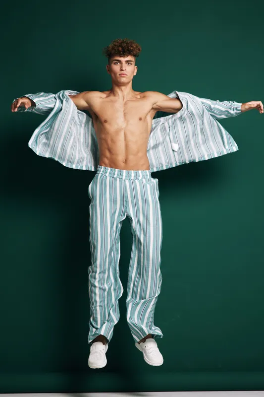 Young man with curly hair wearing striped pajamas and white slippers posing with open shirt and bare chest on dark green background in studio. Fashion, casual and confident style.
