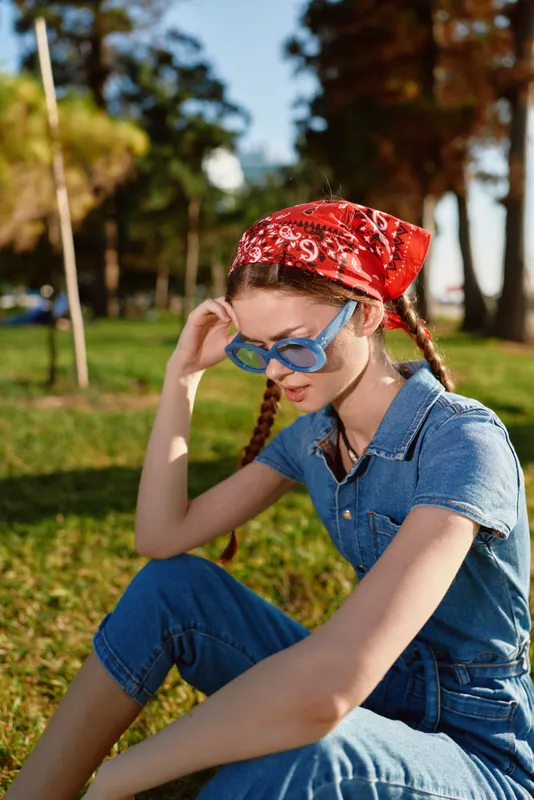 lifestyle woman wearing modern boho-western denim outfit and red bandana, blue sunglasses, sitting outdoors in natural sunny park, casual street style with film color