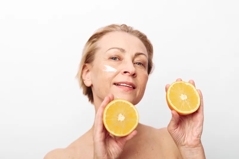Mature woman with short blonde hair holds two orange halves near face with cream on cheek. Concept for natural skincare, healthy lifestyle, beauty routine, and citrus wellness.