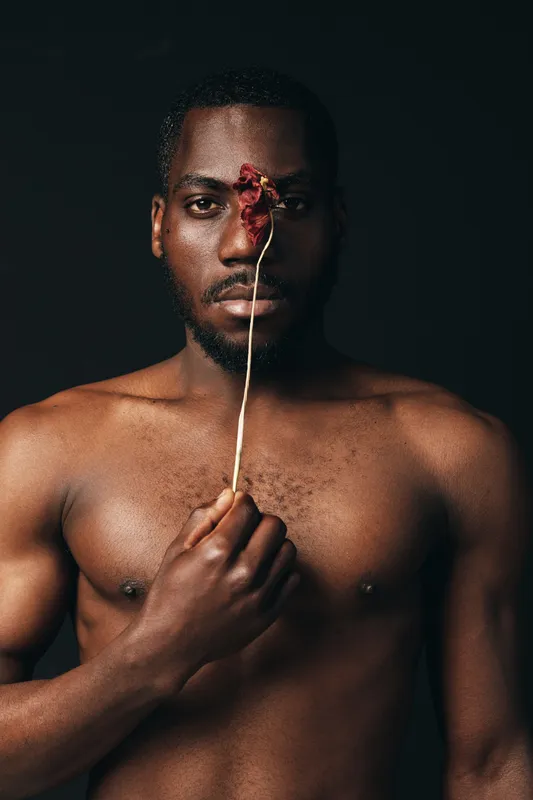 Black man with dark skin and bare chest holding dried flower in front of face in creative visual design style on black background. Artistic portrait with conceptual mood and expression.