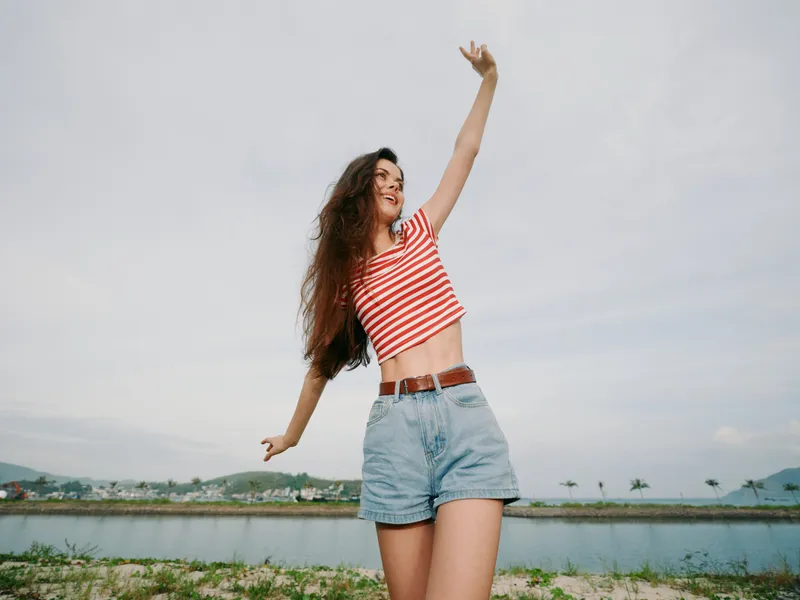 Joyful young woman in a striped crop top and denim shorts stands outdoors by a tranquil waterway, arms raised in celebration under a bright, expansive sky.