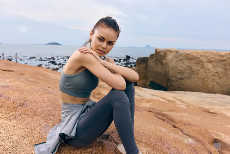 A young woman in activewear sitting on rocks near the ocean, showcasing athletic style and contemplative emotion, perfect for wellness and fitness themes