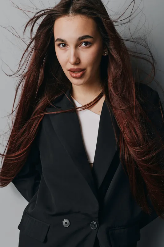 Young woman with long brown hair wearing black blazer and white top posing against gray background with hair flowing, looking confident and stylish in studio portrait photography.