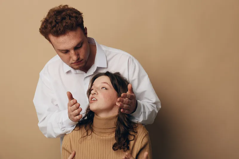 Man in white shirt gently holds woman with curly hair wearing brown sweater. Intimate moment with soft lighting on beige background. Concept of care, support, and connection between couple.