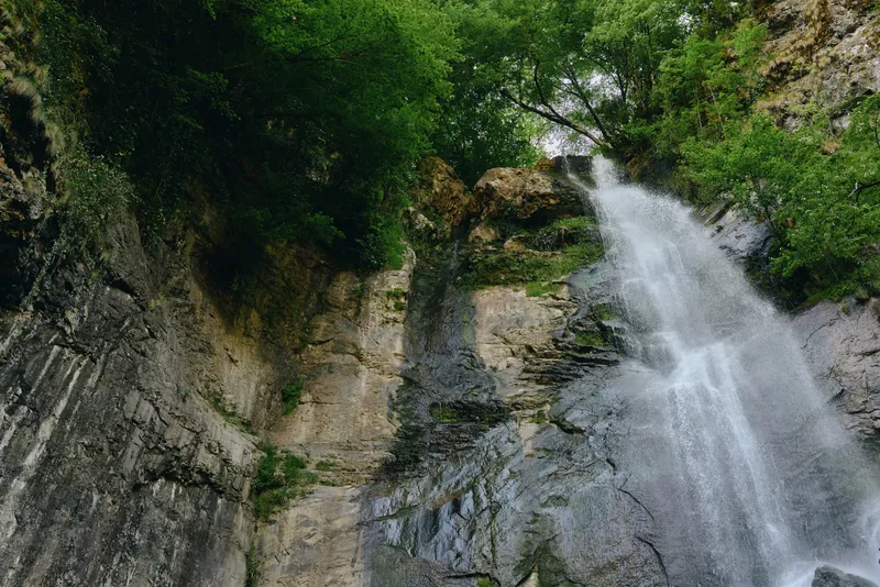 Waterfall nature landscape with lush green foliage and rocky cliffside flowing with clear water under bright daylight in a serene outdoor environment.