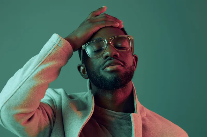 A contemplative Black man in stylish eyewear and a modern jacket, expressing deep thought with a subtle smile against a colorful background