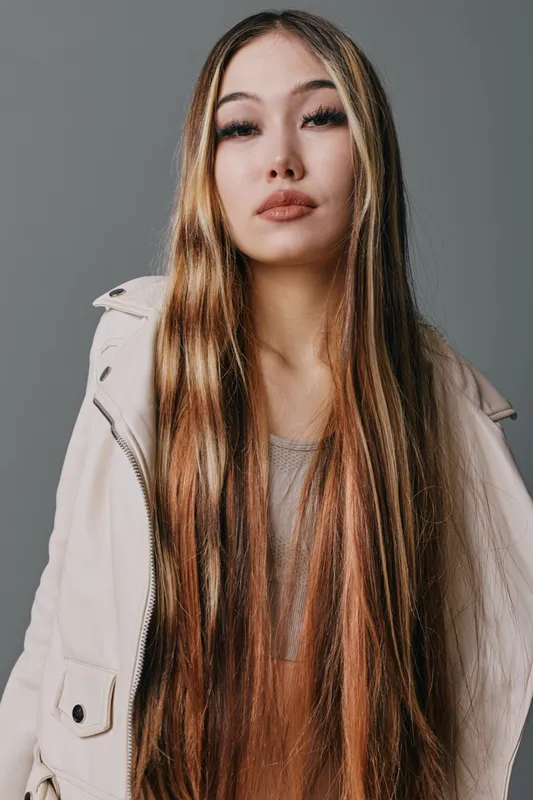 Young woman with long straight hair and neutral expression wearing light jacket posing on gray background for fashion portrait and lifestyle concept.