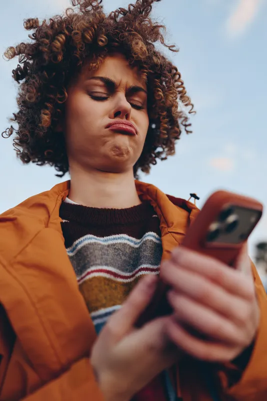 Woman with curly hair holds a smartphone outdoors, casual lifestyle portrait, she looks at the screen with soft light, modern communication moment on a city walk today