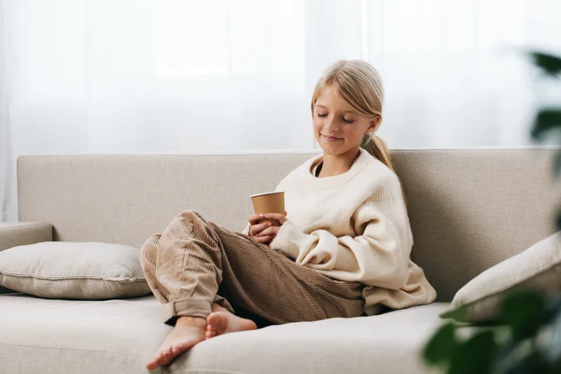 Authentic lifestyle scene of a young girl at home, cozy sweater on a sofa, sipping a cup, a quiet moment after school in soft natural light.