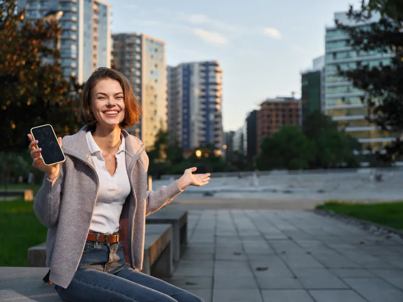 A cheerful woman is seen outdoors in an urban plaza, smiling while holding a smartphone, creating a friendly and approachable moment in a modern city setting.