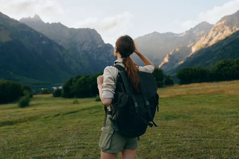 Young woman hiking in mountains, standing on grassy field, wearing backpack, looking at rocky peaks, outdoor adventure, natural daylight, casual outfit