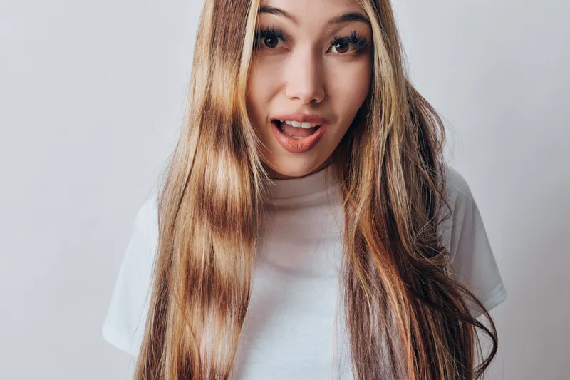 Young woman with long straight hair and open mouth posing on white background. Female model in casual white t shirt with expressive face and natural makeup closeup portrait.